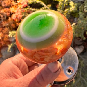 May include: A green and orange glass shift knob with a white ring around the center. The knob is being held by a hand.
