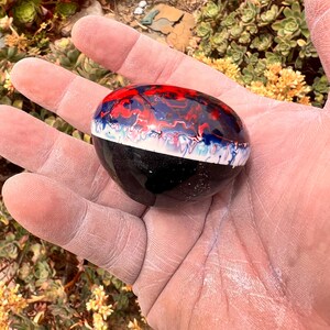 May include: A smooth, rounded stone featuring a glossy finish with a distinct color pattern. The top section displays a marbled blend of red, blue, and white, while the bottom half is solid black, separated by a thin, textured white line.