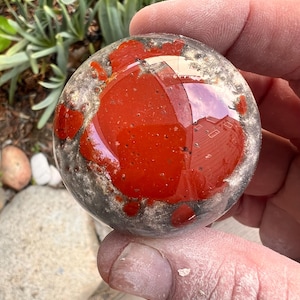 May include: A clear, round glass orb with a vibrant red, speckled interior. The orb has a speckled, gray exterior and reflects the surrounding environment. The object is held in a hand, with a natural outdoor background.