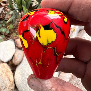May include: A teardrop-shaped glass object featuring a glossy red base color with swirling patterns of bright yellow and black throughout the surface. The object has a smooth, reflective finish and a rounded top that tapers toward a flat base.