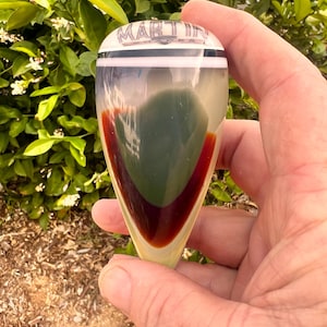 May include: A teardrop-shaped glass bottle stopper with the name "MARTIN" at the top. The stopper features a white band, a green center, and a deep red accent. The stopper is held in a hand against a backdrop of green foliage.