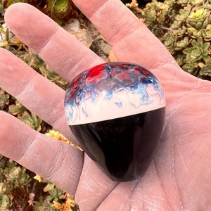 May include: A smooth, rounded gear shift knob featuring a solid black base and a top section with a swirling, marbled pattern of red, white, and blue colors. The object has a glossy, polished finish and a tapered, bulbous shape.