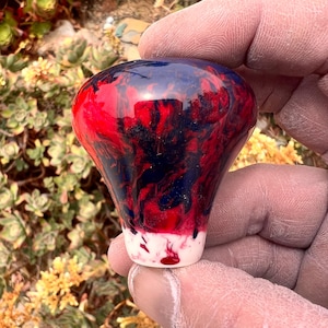 May include: A gear shift knob featuring a glossy, marbled finish with swirling patterns of deep blue, vibrant red, and a solid white base. The object has a rounded, bulbous top that tapers down to a narrower cylindrical bottom section.