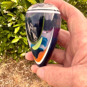 May include: A hand holds a decorative glass bottle stopper. The stopper is teardrop-shaped with a dark blue base, white and blue stripes at the top, and colorful abstract designs in shades of orange, yellow, and blue.