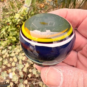 May include: A spherical glass paperweight featuring a smooth, polished surface with horizontal bands of color. The design includes a deep blue base, a thin white stripe, a vibrant yellow ring, and a dark green top section with a clear glass dome.