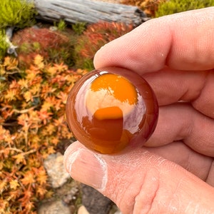 May include: A round, orange agate sphere with a translucent, yellow center. The sphere is smooth and polished, with a high gloss finish.