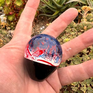 May include: A spherical gear shift knob featuring a smooth, glossy finish. The top half displays a swirling, marbled pattern of deep blue, bright red, and white colors, while the bottom half is a solid, opaque black, creating a distinct contrast.