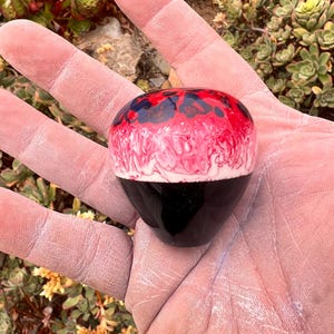 May include: A rounded gear shift knob featuring a glossy finish with a distinct color pattern. The top section displays a marbled blend of vibrant red, white, and dark blue, while the bottom half is a solid, smooth, and polished black surface.