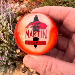 May include: A circular, bright orange resin object featuring a black surfboard silhouette graphic. The design includes a red hand reaching over the surfboard, with the text MARTIN in red and SHAPES in black on a red rectangular background.