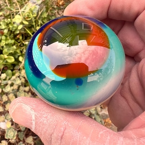 May include: A smooth, spherical glass paperweight featuring a swirling blend of colors including deep blue, vibrant turquoise, and a prominent central patch of burnt orange. The surface is highly reflective, showing distorted outdoor reflections.
