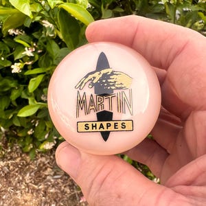 May include: A light pink, round object with the text "MARTIN SHAPES" in black and gold. The design includes a black shape and a gold hand. The object is held in a hand.