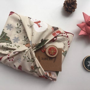 以下が含まれることがあります： クリスマス柄のベージュ色の布で包まれたギフト。結び目があります。 金色の雪の結晶が付いた小さな木製タグと「THANK YOU」カードが添付されています。 松ぼっくりと紙の星も見えます。