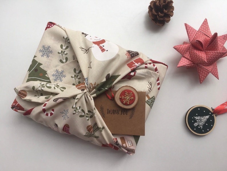 以下が含まれることがあります： クリスマス柄のベージュ色の布で包まれたギフト。結び目があります。 金色の雪の結晶が付いた小さな木製タグと「THANK YOU」カードが添付されています。 松ぼっくりと紙の星も見えます。