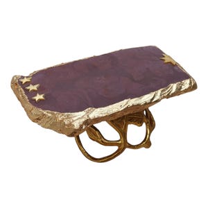 Op de afbeelding: Een statement ring met een rechthoekige, dieppaarse stenen plaat met een getextureerde rand van bladgoud. Het oppervlak is versierd met drie kleine gouden sterren aan de linkerkant en één aan de rechterkant. De ringband is een goudkleurige takstructuur.
