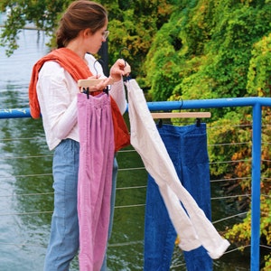 May include: A selection of pants in various colors, including a pair of light pink corduroy pants, a pair of cream-colored pants, and a pair of blue denim pants. The pants are hanging on wooden hangers. The background features lush green foliage and a body of water.