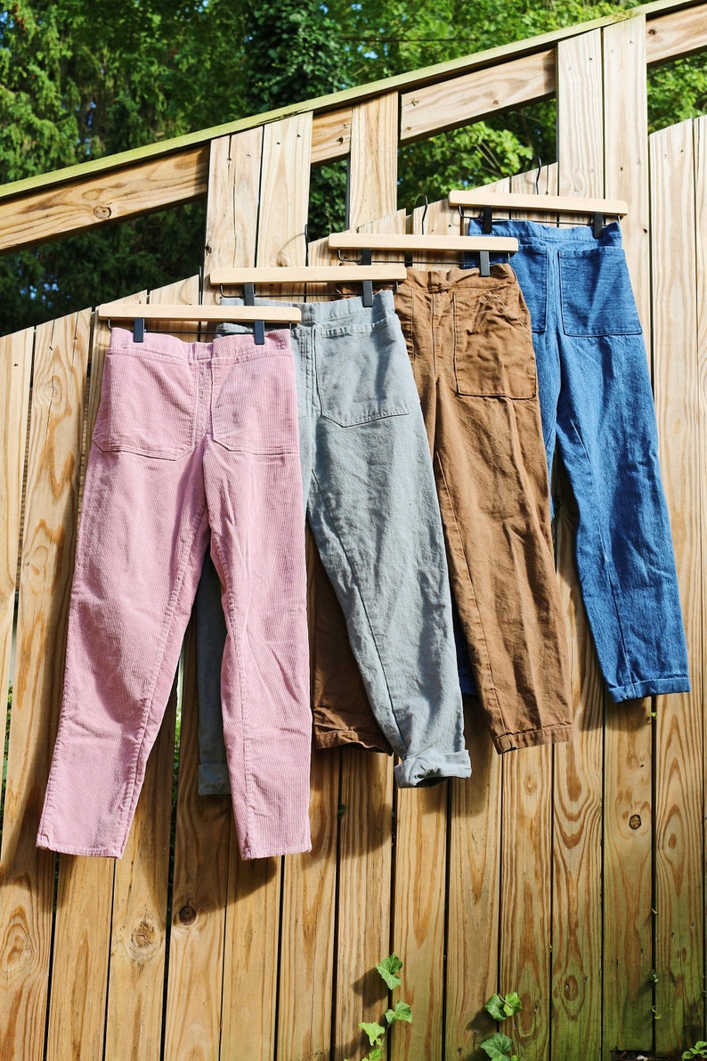 May include: Four pairs of pants in various colors hang on a wooden fence. The colors include pink, gray, brown, and blue. Each pair of pants has a pocket on the back and a front pocket. The pants are made of a soft material.