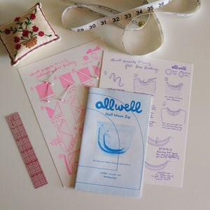 May include: A set of sewing instructions for a half moon zip, with a pink and white instruction sheet, a blue and white instruction booklet, and a pink ruler. The instruction sheet has the text "allwell presents" and "flat for finding". The instruction booklet has the text "allwell" and "Half Moon Zip".
