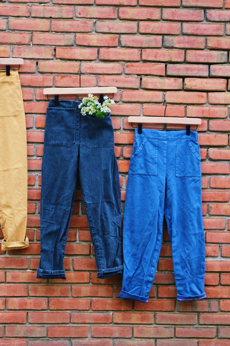May include: Three pairs of pants hang against a red brick wall. The pants are in shades of yellow, dark blue, and light blue. The dark blue pants have a pocket filled with white flowers. All pants have a cuffed hem.