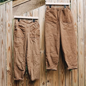 May include: Two pairs of brown pants hanging on a wooden fence. The pants have large front pockets and a relaxed fit. One pair has a high waist and elastic waistband. The pants are made of a sturdy fabric.