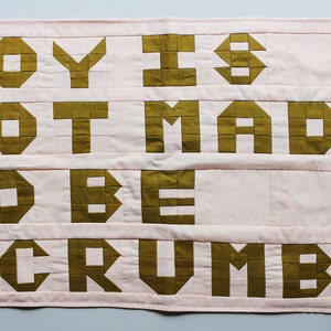 May include: A pink fabric banner with the text "JOY IS NOT MADE TO BE A CRUMB" in gold block letters.