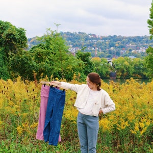 May include: A woman holds up a wooden hanger with two pairs of pants: one pink and one blue. She is standing in a field of yellow flowers, wearing a white jacket and blue pants. A city is visible in the background.