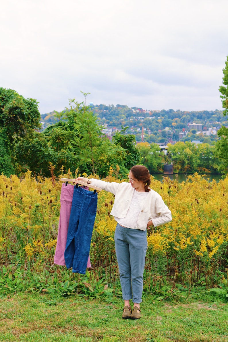 May include: A woman holds up a wooden hanger with two pairs of pants: one pink and one blue. She is standing in a field of yellow flowers, wearing a white jacket and blue pants. A city is visible in the background.