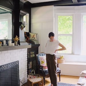 May include: A person wearing a white top and brown pants stands in a room with a fireplace and a blue rug. The room has white walls, black trim, and a large window. A small wooden chair and a basket are also visible.