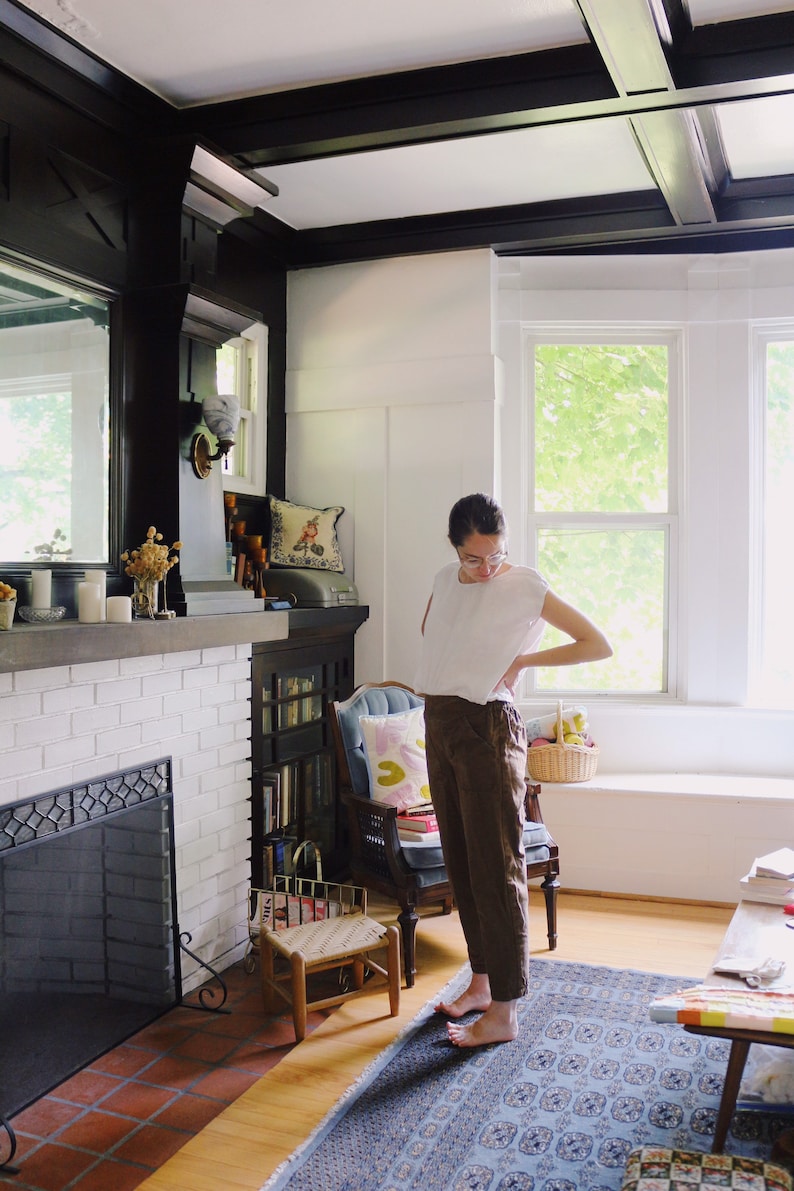 May include: A person wearing a white top and brown pants stands in a room with a fireplace and a blue rug. The room has white walls, black trim, and a large window. A small wooden chair and a basket are also visible.