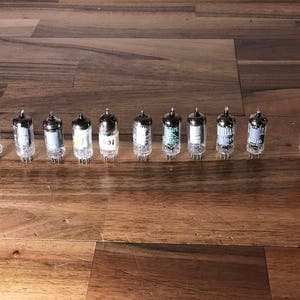 Puede incluir: Una fila de 12 tubos de vacío plateados con varias marcas y etiquetas. Los tubos están dispuestos en línea sobre una superficie de madera.