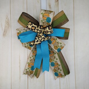 Può includere: Un fiocco blu, marrone e beige con un accento a stampa leopardata. Il fiocco è decorato con un motivo di zucche e foglie.