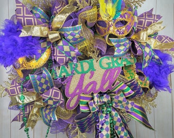 May include: Purple, green, and gold Mardi Gras wreath with a sign that says "Mardi Gras Y'all". The wreath is decorated with feathers, beads, and masks.