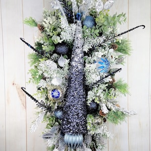 Op de afbeelding: Een blauwe en zilveren kerstkrans met een glinsterende blauwe dennenappel, zilveren en blauwe kerstballen en bevroren groen.