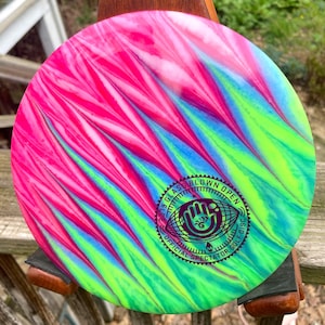 May include: A pink, blue and green disc golf disc with a purple and white logo that says "2019 Glass Blown Open Official Spectator Golf Disc".