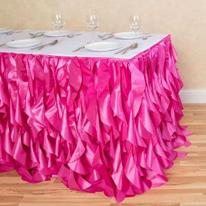 May include: A rectangular table covered with a white tablecloth and a vibrant pink ruffled skirt. The table is set with white plates, silverware, and wine glasses, suggesting a formal dining setting. The ruffled skirt adds a decorative touch.