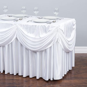 May include: A white rectangular table covered with a white satin tablecloth and a pleated skirt. The table is set with white plates, silverware, and wine glasses. The table is on a dark wood floor.