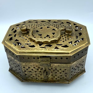 May include: An antique brass octagonal box with intricate openwork and a hinged lid. The box features a decorative handle and a latch closure. The brass has a warm, aged patina, suggesting its vintage origin. The design includes floral and geometric patterns.