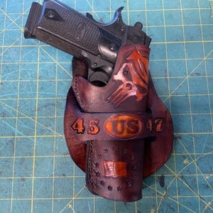 Può includere: Una pistola nera in una fondina di cuoio marrone. La fondina presenta un disegno a teschio, la scritta "45 US 47" e una bandiera americana. La fondina è realizzata in pelle marrone scuro con accenti arancioni.