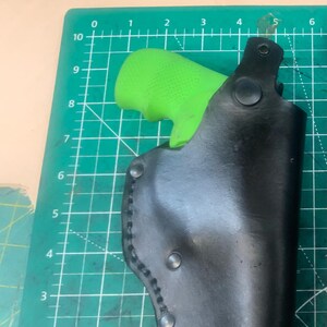 May include: A black leather holster with a green grip inside. The holster is secured with rivets and stitching. The background is a green cutting mat with grid lines.