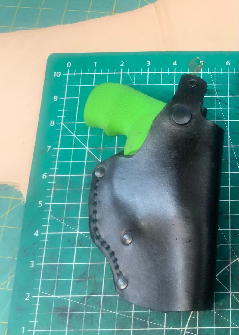May include: A black leather holster with a green grip inside. The holster is secured with rivets and stitching. The background is a green cutting mat with grid lines.