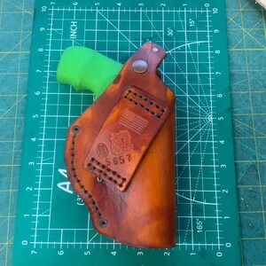 May include: A brown leather holster with a stitched design and a small American flag emblem. The holster is shown on a green cutting mat with inch measurements. The holster is designed to hold a handgun.