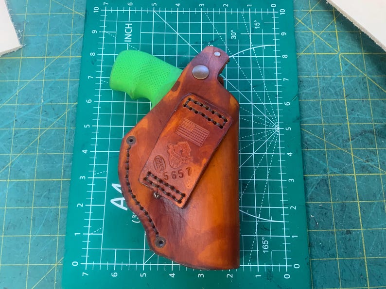 May include: A brown leather holster with a stitched design and a small American flag emblem. The holster is shown on a green cutting mat with inch measurements. The holster is designed to hold a handgun.