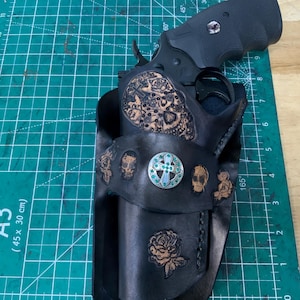 May include: A black leather holster containing a black revolver. The holster features embossed skull and rose designs, with a central silver-toned circular concho ornament inlaid with small green stones. The leather is stitched with black thread.
