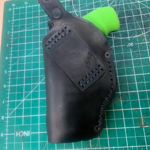 May include: Black leather holster with a green grip visible. The holster has a stitched border and a rectangular patch with an emblem and the number 4552. The holster is on a green cutting mat.