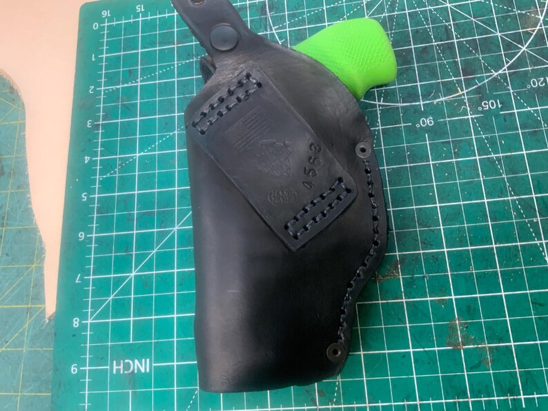 May include: Black leather holster with a green grip visible. The holster has a stitched border and a rectangular patch with an emblem and the number 4552. The holster is on a green cutting mat.