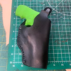 May include: A black leather holster with a bright green handgun inside. The holster is stitched along the edges and has a snap closure. The holster is on a green cutting mat.