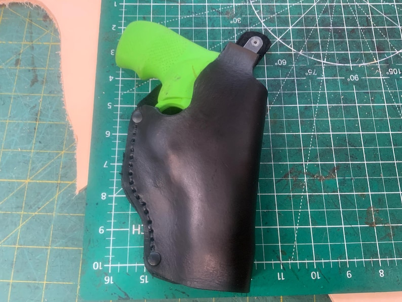 May include: A black leather holster with a bright green handgun inside. The holster is stitched along the edges and has a snap closure. The holster is on a green cutting mat.