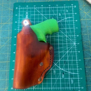 May include: A brown leather holster with a green grip pistol inside. The holster is secured with metal rivets and stitching. The holster is on a green cutting mat with inch measurements.