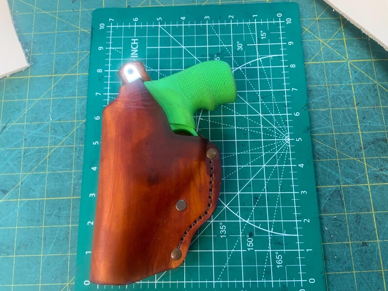 May include: A brown leather holster with a green grip pistol inside. The holster is secured with metal rivets and stitching. The holster is on a green cutting mat with inch measurements.