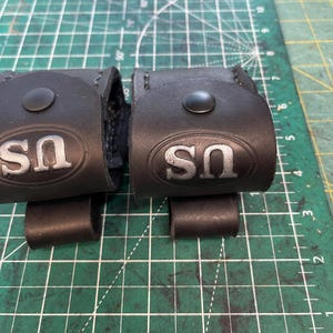 May include: Two black leather pouches featuring a flap closure with a black metal snap button. Each pouch is embossed with the letters US inside an oval frame. The pouches are constructed with thick black stitching and include a belt loop on the back.