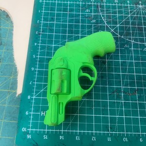May include: A bright green toy revolver, resting on a green cutting mat with grid lines and degree markings. The toy gun is a detailed replica, featuring a handle, trigger, and cylinder. The background includes a piece of beige material.